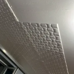 Fragment sufitu z zamontowanymi białymi panelami 3D o wzorze rombów, widoczne okno w tle, oświetlenie górne.