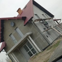 SpecINSTAL Adrian Strugała - Dwupiętrowy dom w trakcie malowania elewacji, widoczny zza szyby samochodu, z rusztowaniem i pracownikiem w kombinezonie ochronnym.