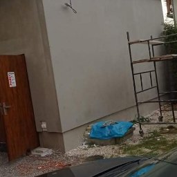 SpecINSTAL Adrian Strugała - Świeżo otynkowana elewacja budynku z drewnianymi drzwiami i metalowym rusztowaniem, widok zza szyby samochodu.