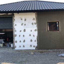 SpecINSTAL Adrian Strugała - Elewacja budynku w trakcie ocieplania w Wadowicach Górnych: fragment ściany z przyklejonym styropianem, fragment otynkowany na szaro, otwarty garaż z widocznym wnętrzem i rusztowania.