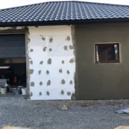 Elewacja budynku w trakcie ocieplania w Wadowicach Górnych: fragment ściany z przyklejonym styropianem, fragment otynkowany na szaro, otwarty garaż z widocznym wnętrzem i rusztowania.