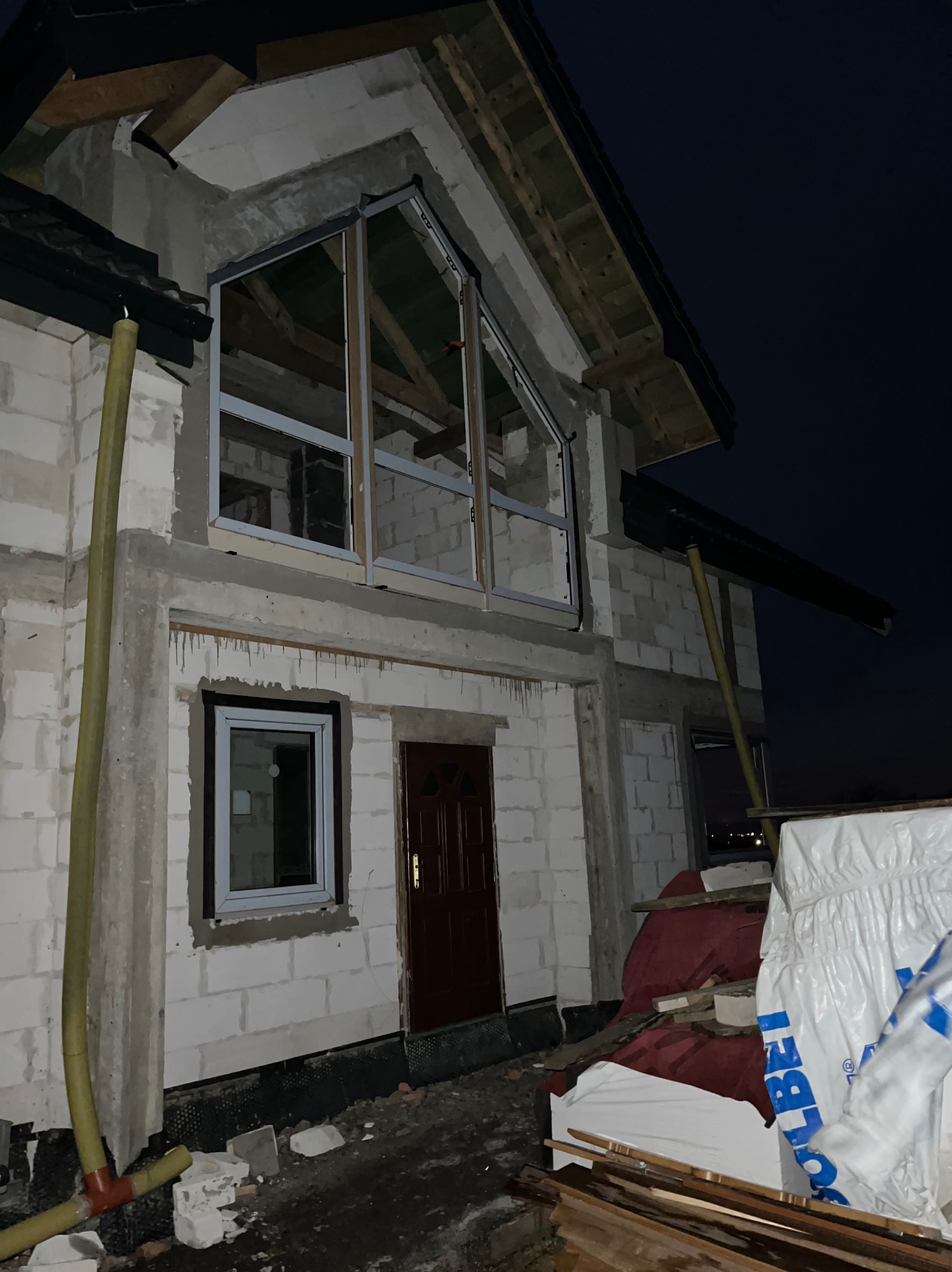 Budynek w stanie surowym otwartym z zamontowanymi oknami i drzwiami wejściowymi, widoczne pustaki, niezabezpieczone drewniane elementy konstrukcyjne dachu i rury spustowe, nocne ujęcie.