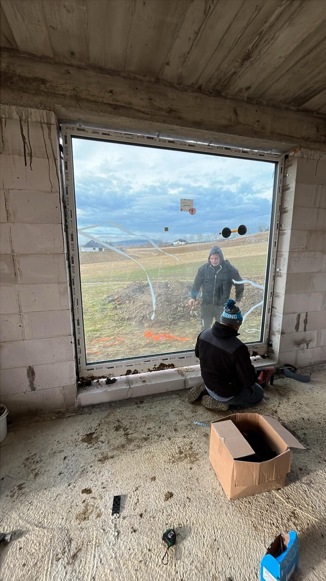 Dwóch mężczyzn instaluje duże okno w surowym stanie, widoczne przyssawki do szkła, narzędzia i karton na podłodze.