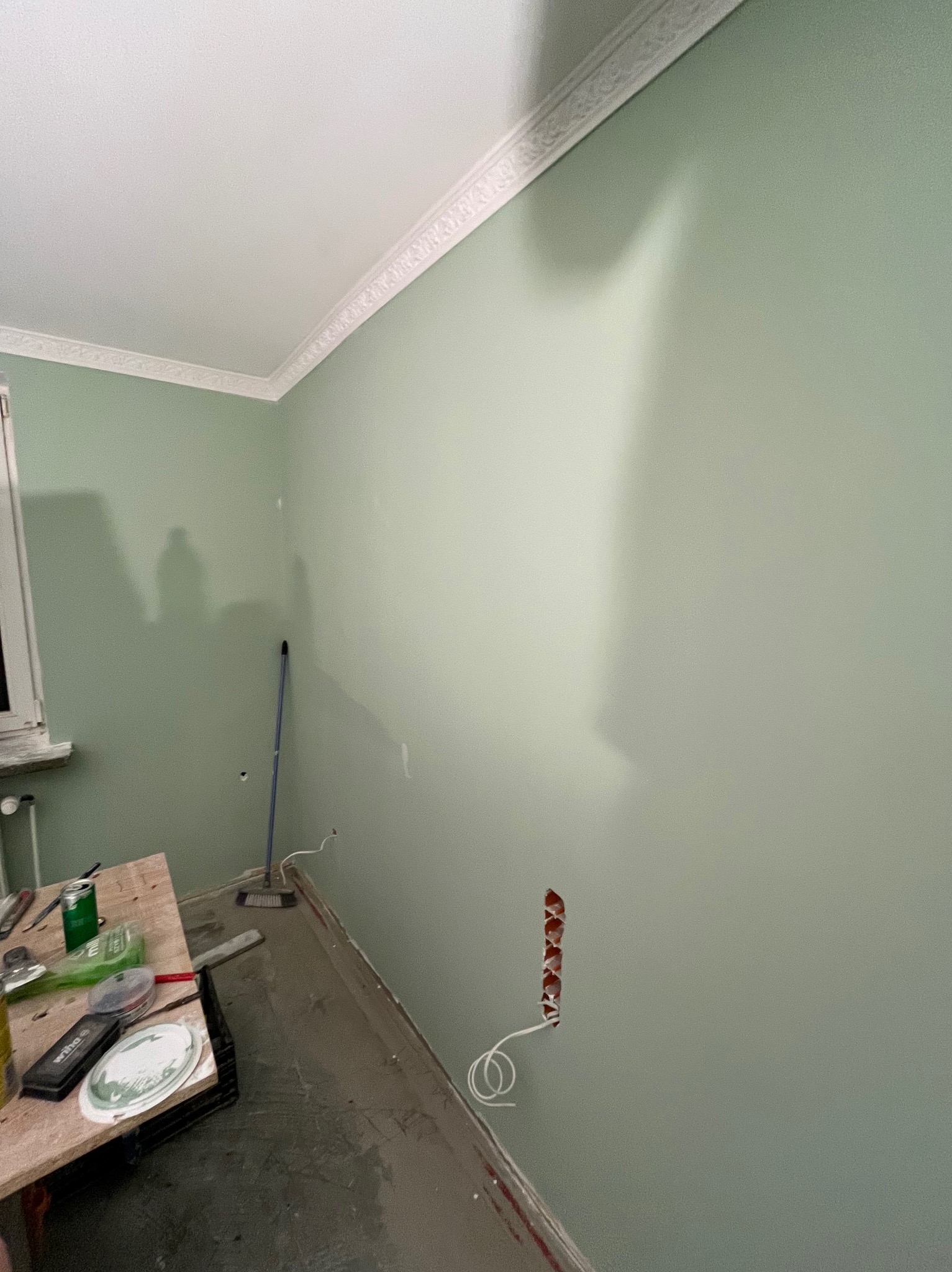Świeżo pomalowane, jasnozielone ściany w pokoju z ozdobną listwą przysufitową. Widoczne gniazdko elektryczne bez osłonki, narzędzia malarskie na prowizorycznym stole oraz ślady po remoncie...