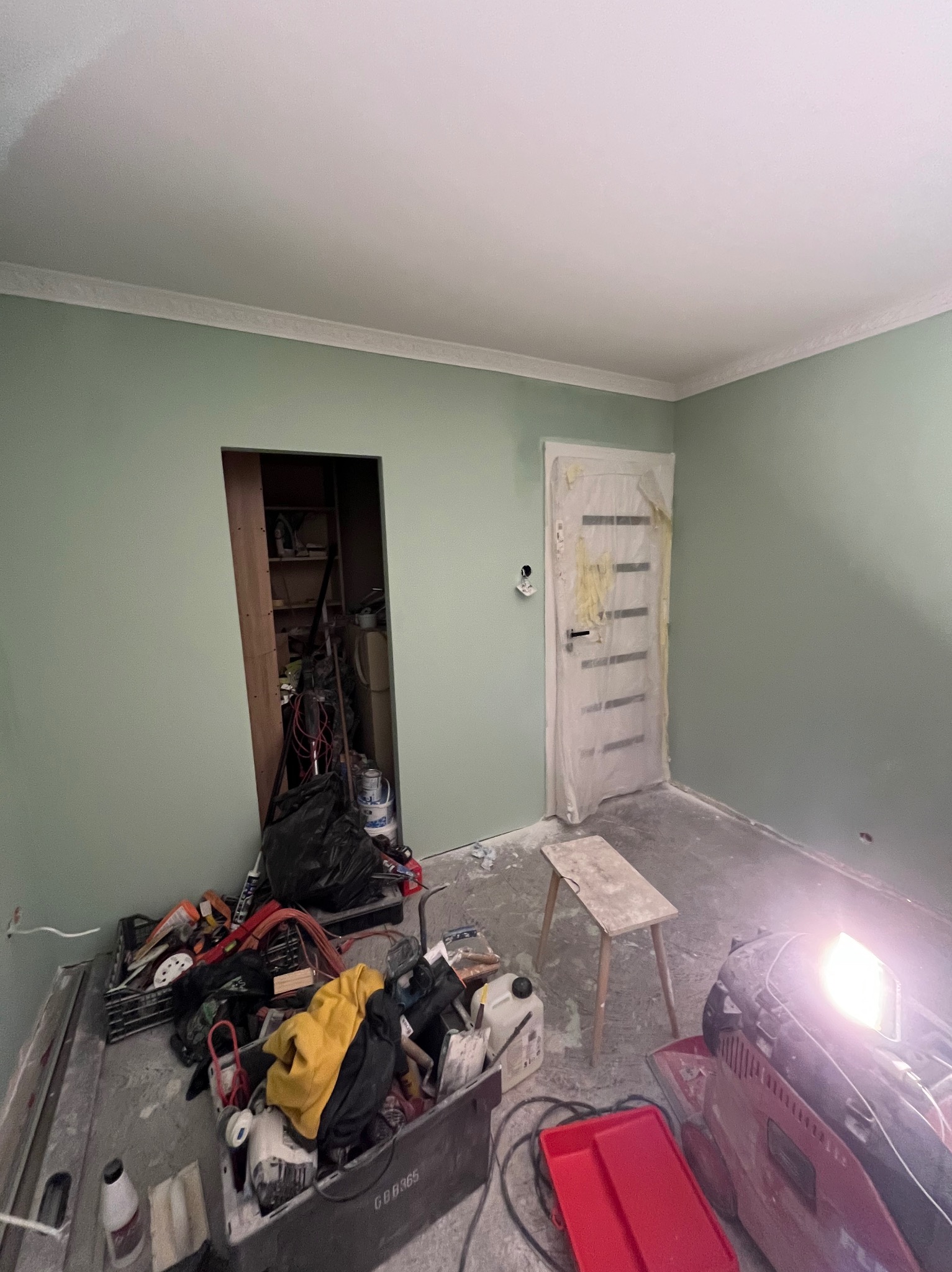 Pokój w trakcie remontu: świeżo pomalowane ściany na pistacjowy kolor, zabezpieczone drzwi folią malarską, widoczne narzędzia i akcesoria malarskie oraz prowizoryczny stołek.