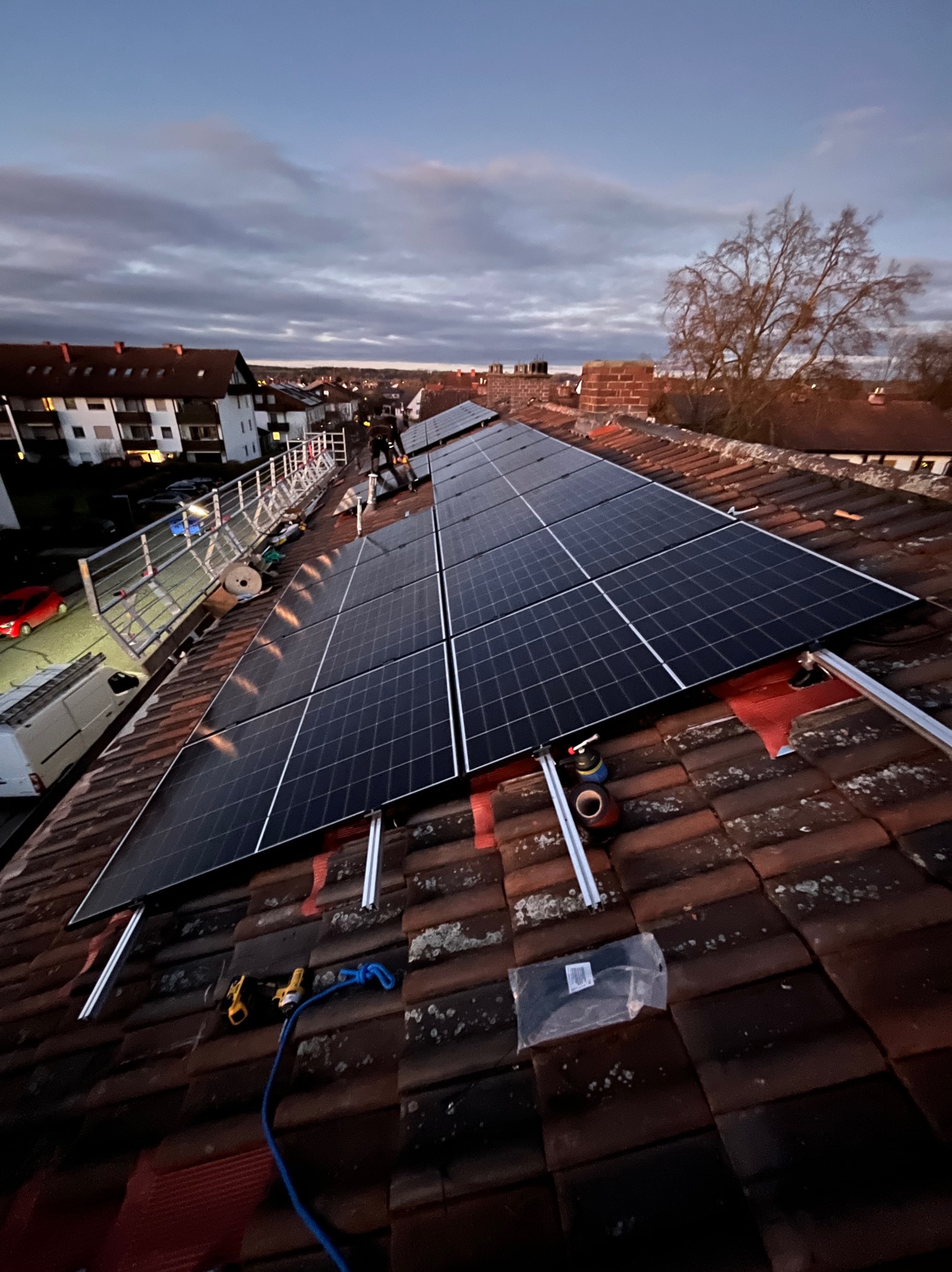 Panele słoneczne w trakcie montażu na dachu z czerwonej dachówki, widoczne narzędzia i zabezpieczenia, rusztowanie w tle, pochmurne niebo.