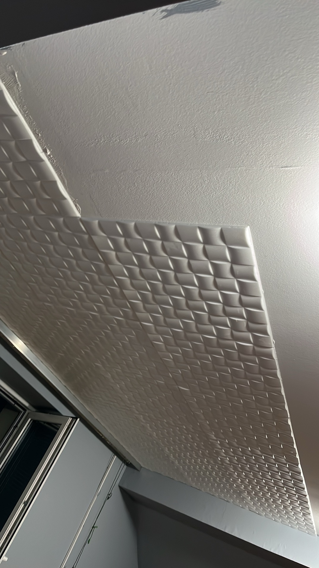 Fragment sufitu z zamontowanymi białymi panelami 3D o wzorze rombów, widoczne okno w tle, oświetlenie górne.