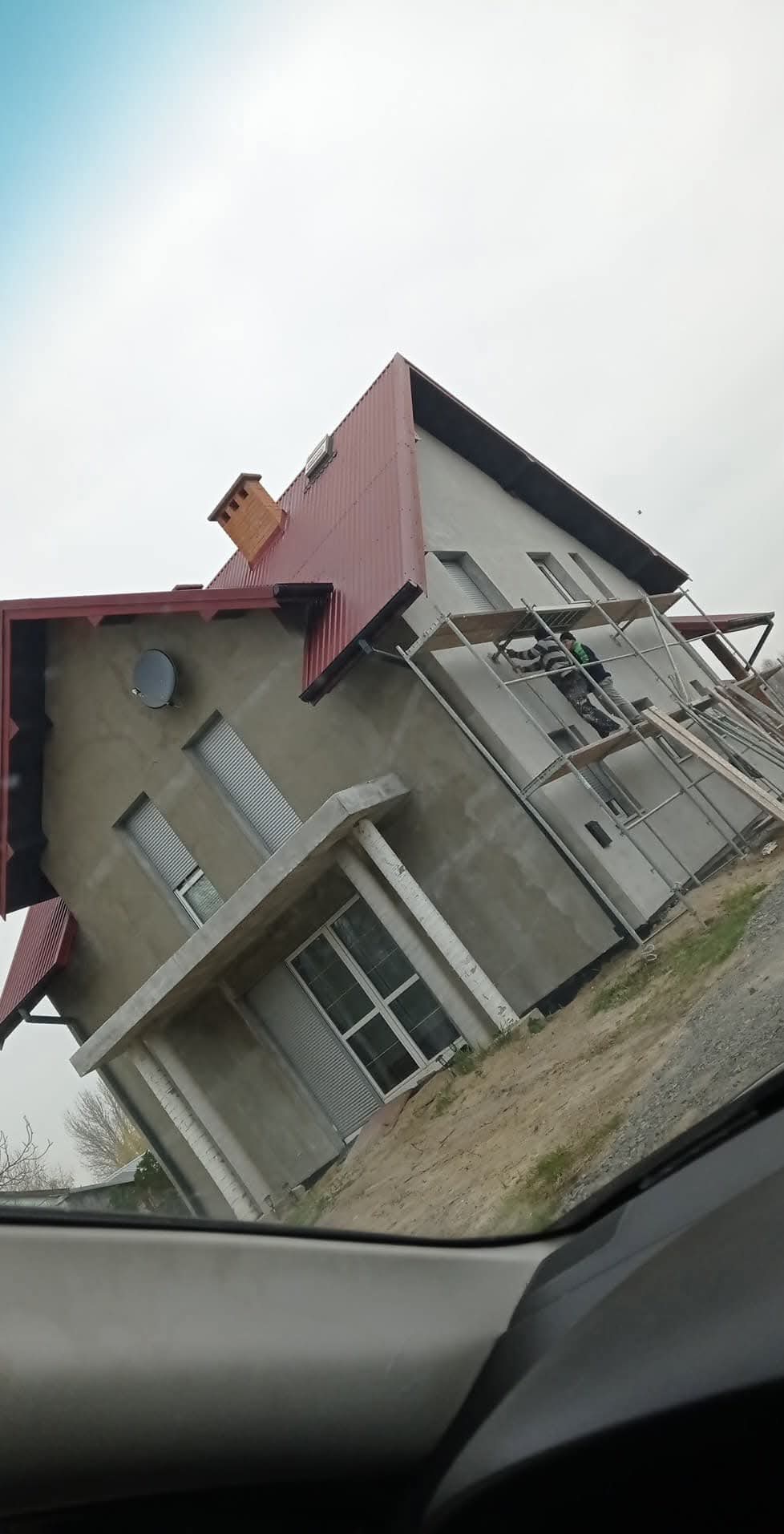 Dwupiętrowy dom w trakcie malowania elewacji, widoczny zza szyby samochodu, z rusztowaniem i pracownikiem w kombinezonie ochronnym.