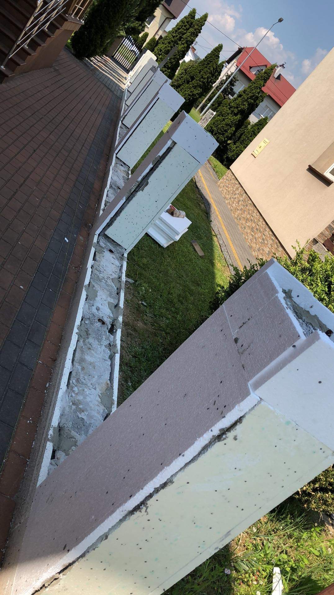 Fragment elewacji budynku w trakcie ocieplania, widoczne płyty styropianowe, warstwa kleju i siatki, przygotowane do dalszych prac wykończeniowych, z widocznym fragmentem chodnika i trawnika w tle.
