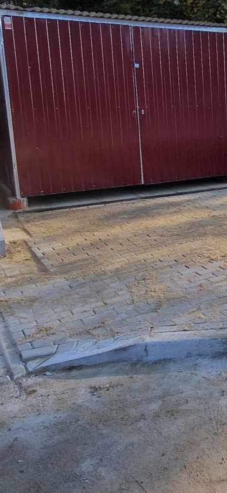 Ciemnoczerwona brama garażowa z pionowymi panelami, widoczna z poziomu gruntu, z kostką brukową przed nią.