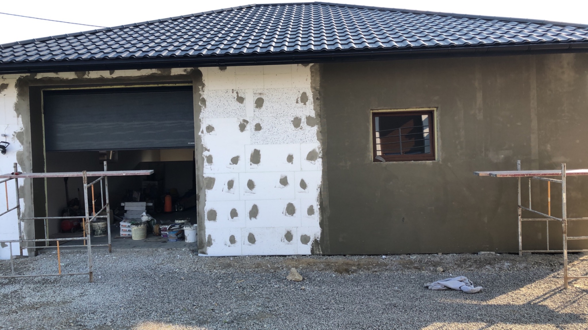 Elewacja budynku w trakcie ocieplania w Wadowicach Górnych: fragment ściany z przyklejonym styropianem, fragment otynkowany na szaro, otwarty garaż z widocznym wnętrzem i rusztowania.