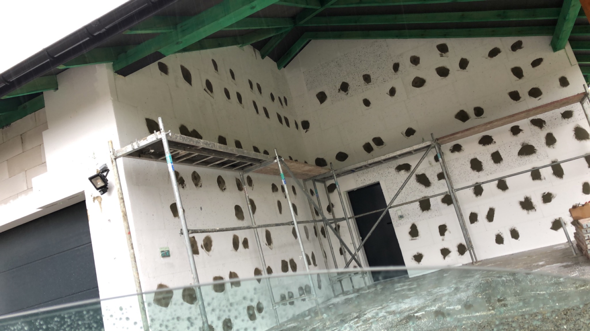 Przygotowanie ściany z bloków betonowych do dalszych prac wykończeniowych, z widocznymi otworami wypełnionymi zaprawą, rusztowaniem i zieloną konstrukcją dachu.