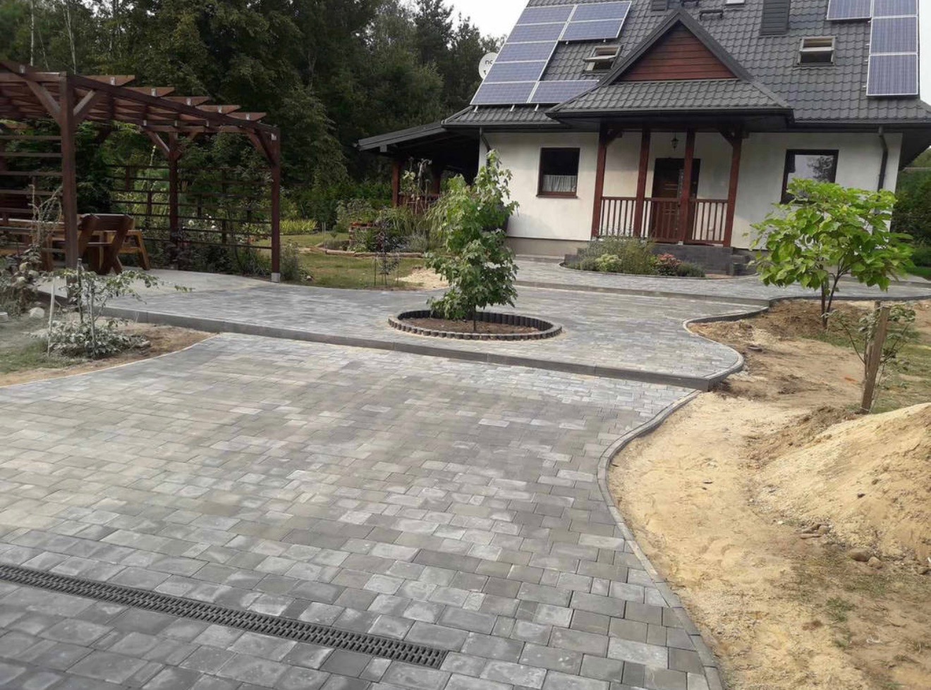 Ułożona szara kostka brukowa tworzy ścieżkę do domu z panelami słonecznymi. Widoczna studzienka kanalizacyjna, pergola i małe drzewko w okręgu z obrzeży.