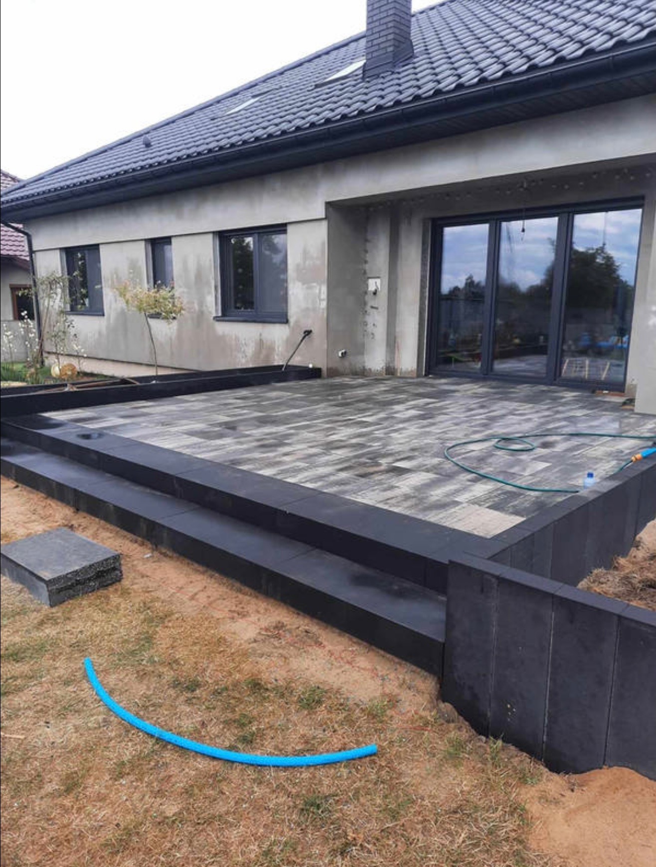 Wykończony taras z kostki brukowej w nowoczesnym domu. Schody z ciemnej kostki prowadzą na taras z szaro-białej kostki. W tle widoczny budynek z dużymi oknami.