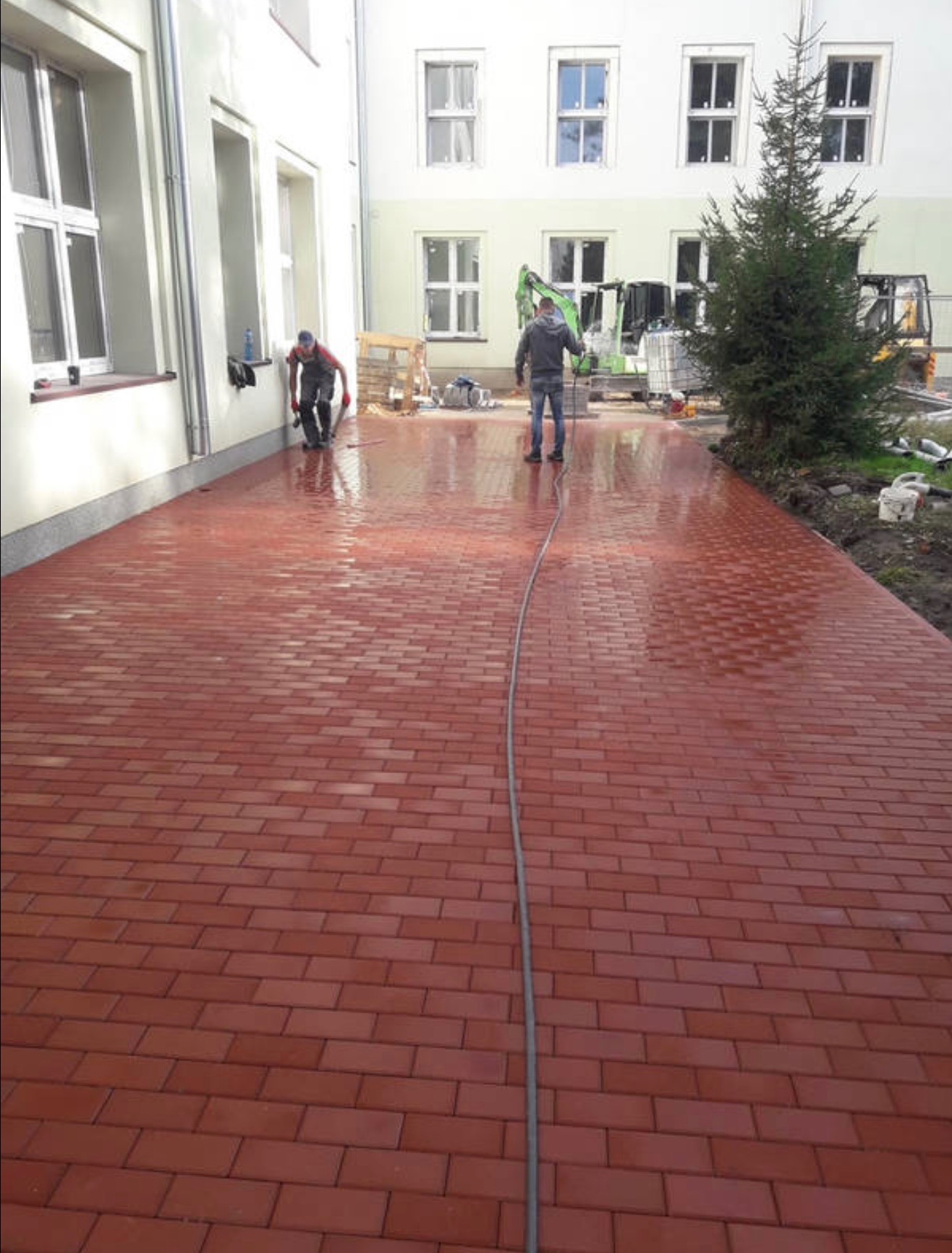 Czerwona kostka brukowa, myta przez dwóch robotników przy budynku z oknami. Na dalszym planie minikoparka i choinka.