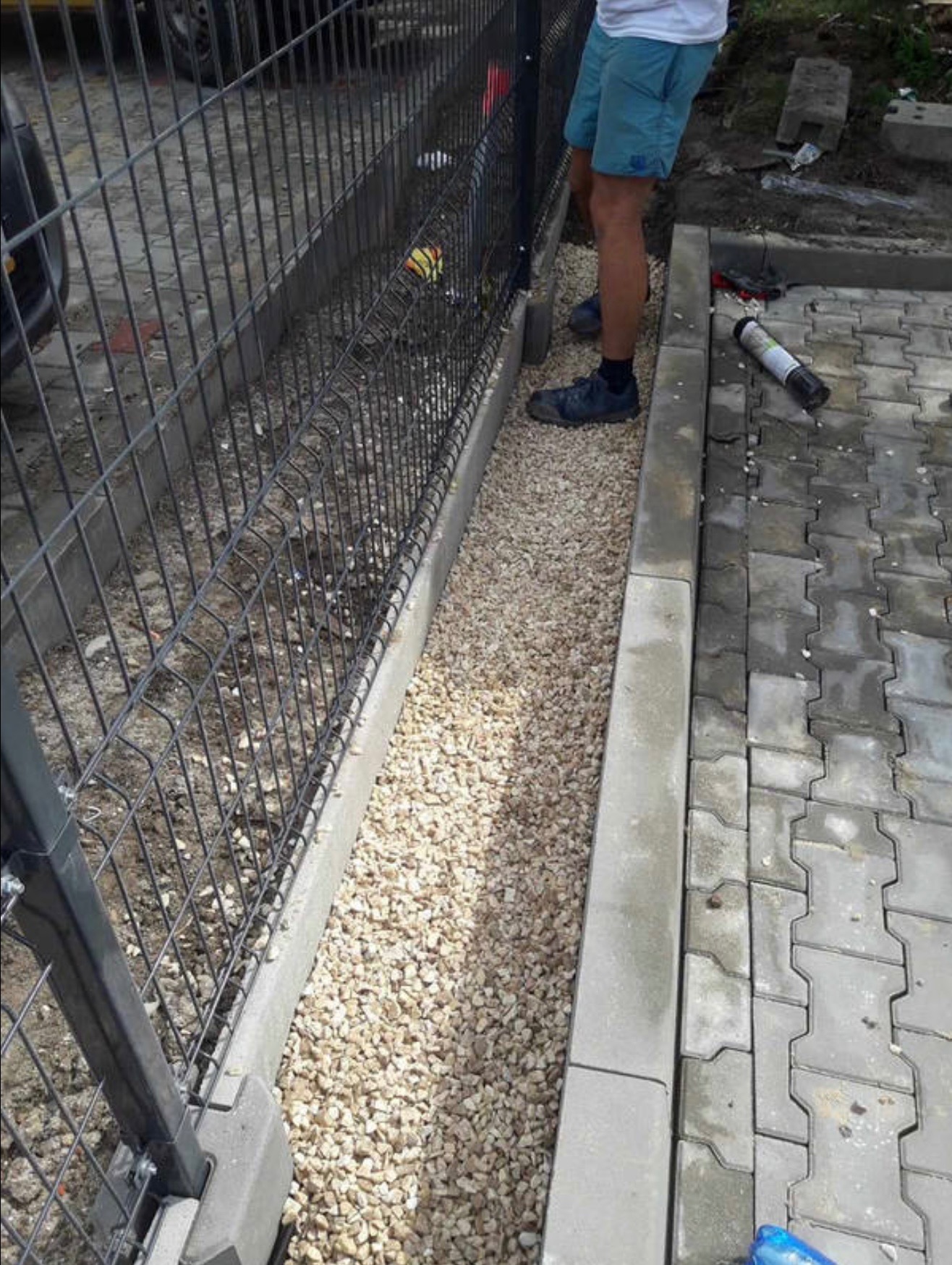 Montaż ogrodzenia panelowego z podsypką żwirową i obrzeżem betonowym. Widoczne fragmenty kostki brukowej i narzędzia montera.