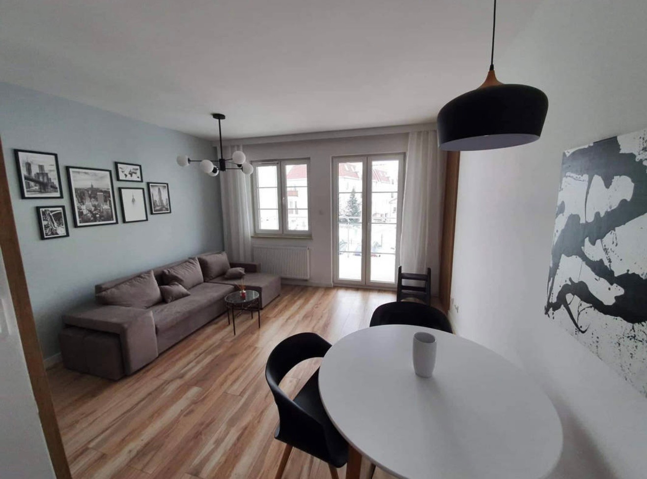 Jasne wnętrze mieszkania z drewnianą podłogą, szarą sofą i białym stołem. Na ścianie wiszą czarno-białe zdjęcia i abstrakcyjny obraz. Widoczne okna z białymi firanami.