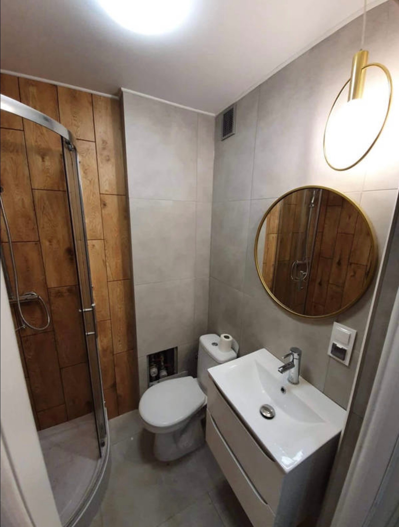 Nowoczesna łazienka z kabiną prysznicową z drewnianą ścianą, okrągłym lustrem w złotej ramie nad białą szafką z umywalką i designerską lampą. Szare płytki na ścianach i podłodze.