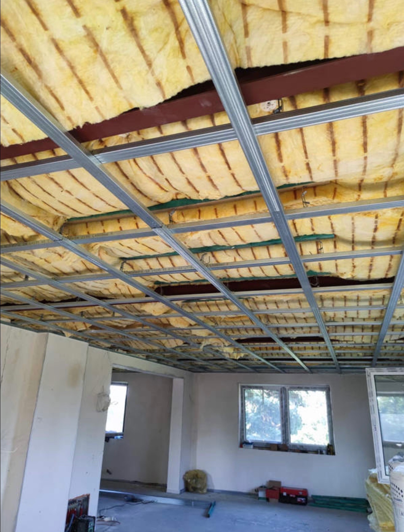 Sufit podwieszany z izolacją z wełny mineralnej i metalową konstrukcją w surowym wnętrzu. Widoczne okna i ściany w trakcie wykończenia.