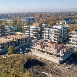 PROPERTY SPÓŁKA Z OGRANICZONĄ ODPOWIEDZIALNOŚCIĄ - Wysoki kąt widokowy na plac budowy z kilkoma budynkami apartamentowymi w różnych stadiach budowy, otoczone zielenią i zabudową mieszkalną w tle, w słoneczny dzień.