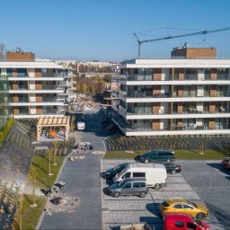 PROPERTY SPÓŁKA Z OGRANICZONĄ ODPOWIEDZIALNOŚCIĄ - Nowoczesne apartamenty z balkonami, widok z góry na parking i teren zagospodarowany zielenią, w tle budowa i dźwig