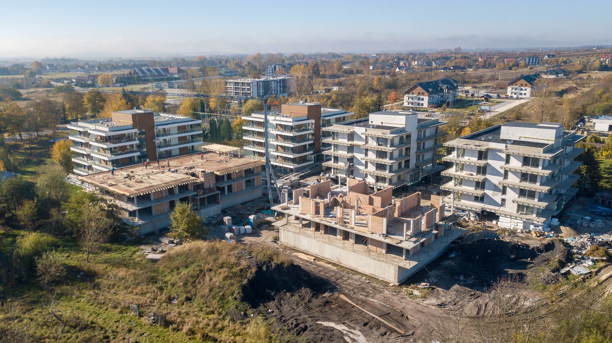 Wysoki kąt widokowy na plac budowy z kilkoma budynkami apartamentowymi w różnych stadiach budowy, otoczone zielenią i zabudową mieszkalną w tle, w słoneczny dzień.
