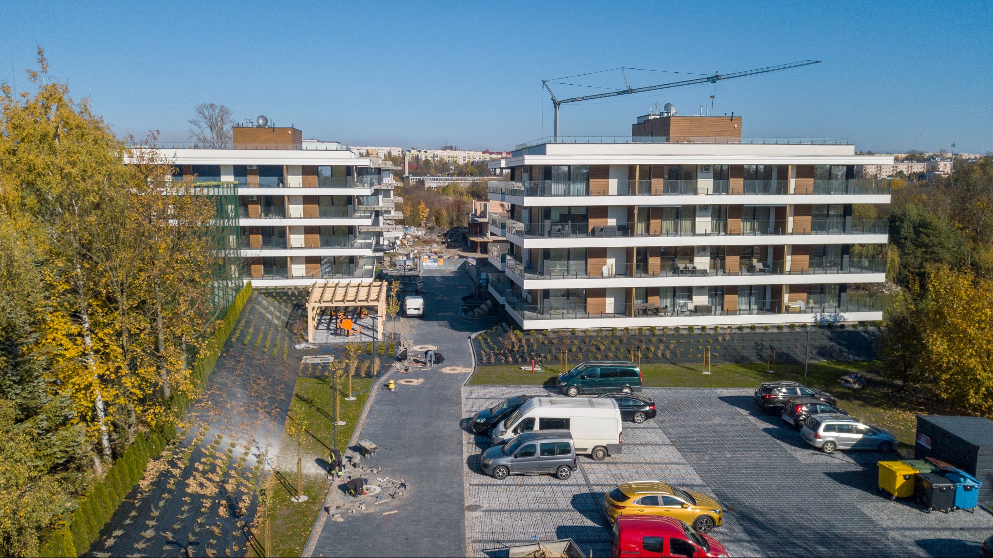 Nowoczesne apartamenty z balkonami, widok z góry na parking i teren zagospodarowany zielenią, w tle budowa i dźwig