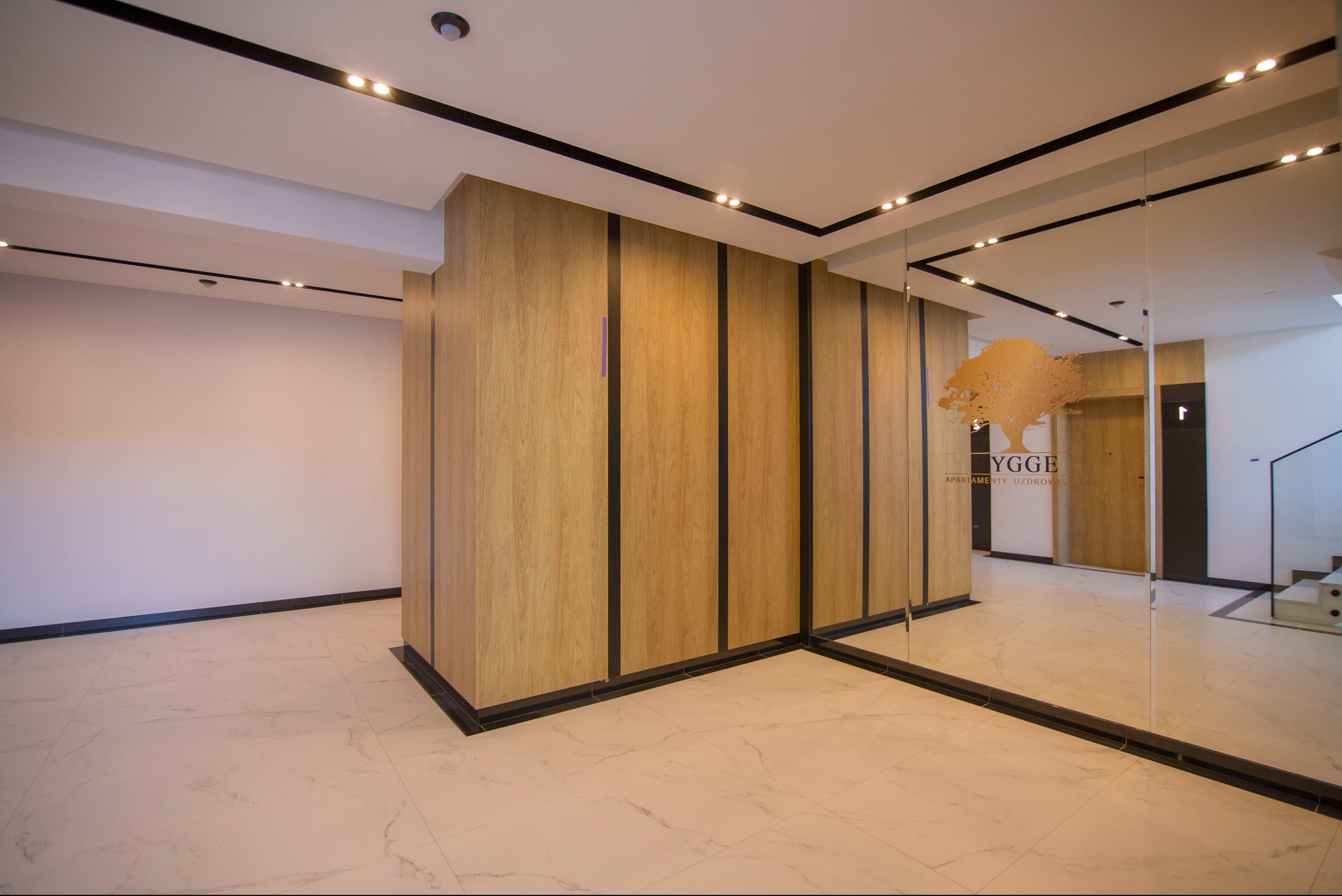 Nowoczesny hol wejściowy z drewnianą zabudową, marmurową podłogą i szklaną ścianą z logo 'HYGGE Apartamenty Uzdrowiskowe'.