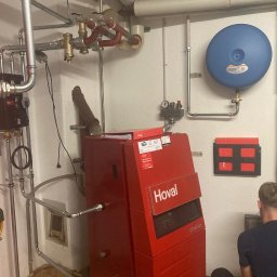 ECO ANGELS SOLUTIONS SPÓŁKA Z OGRANICZONĄ ODPOWIEDZIALNOŚCIĄ - Czerwony piec CO marki Hoval z podłączoną instalacją, widoczny pracownik podczas montażu. Elementy instalacji grzewczej na ścianie i pod sufitem.