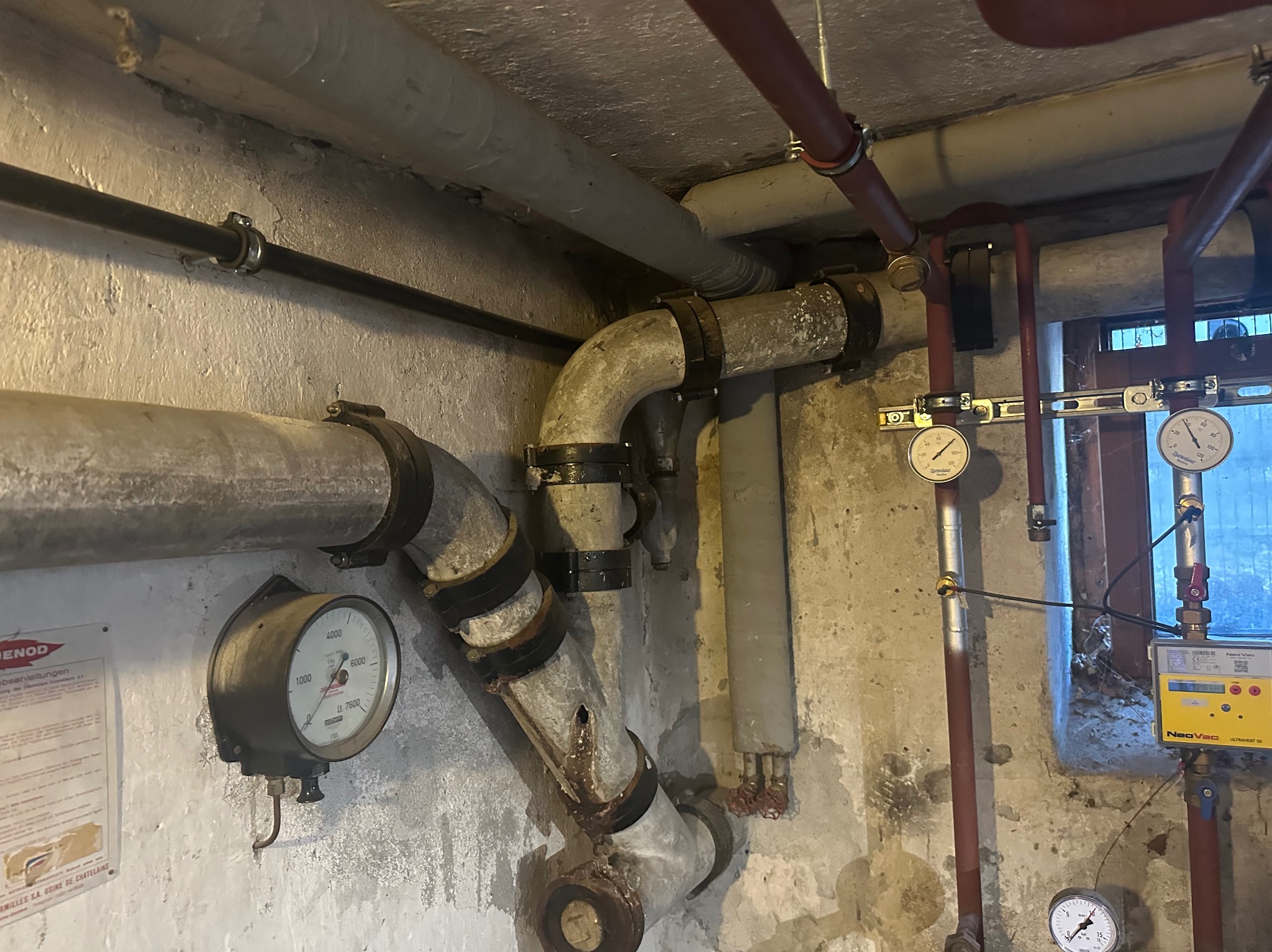 Złożona instalacja rurowa w piwnicy, widoczne rury stalowe szare i czerwone, manometry, zawory, elementy mocujące, ściany z widocznymi śladami wilgoci i zabrudzeń, okno w tle.