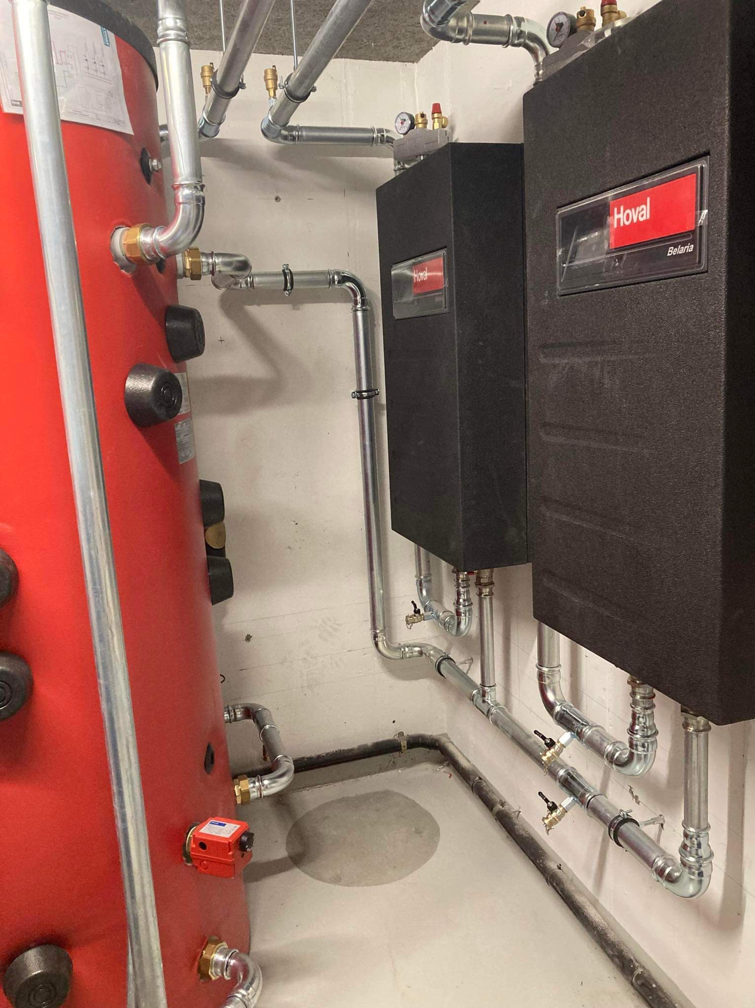 Czerwony zbiornik ciepłej wody użytkowej i dwa czarne kotły grzewcze Hoval Belaria z kompletną instalacją rur w piwnicy.