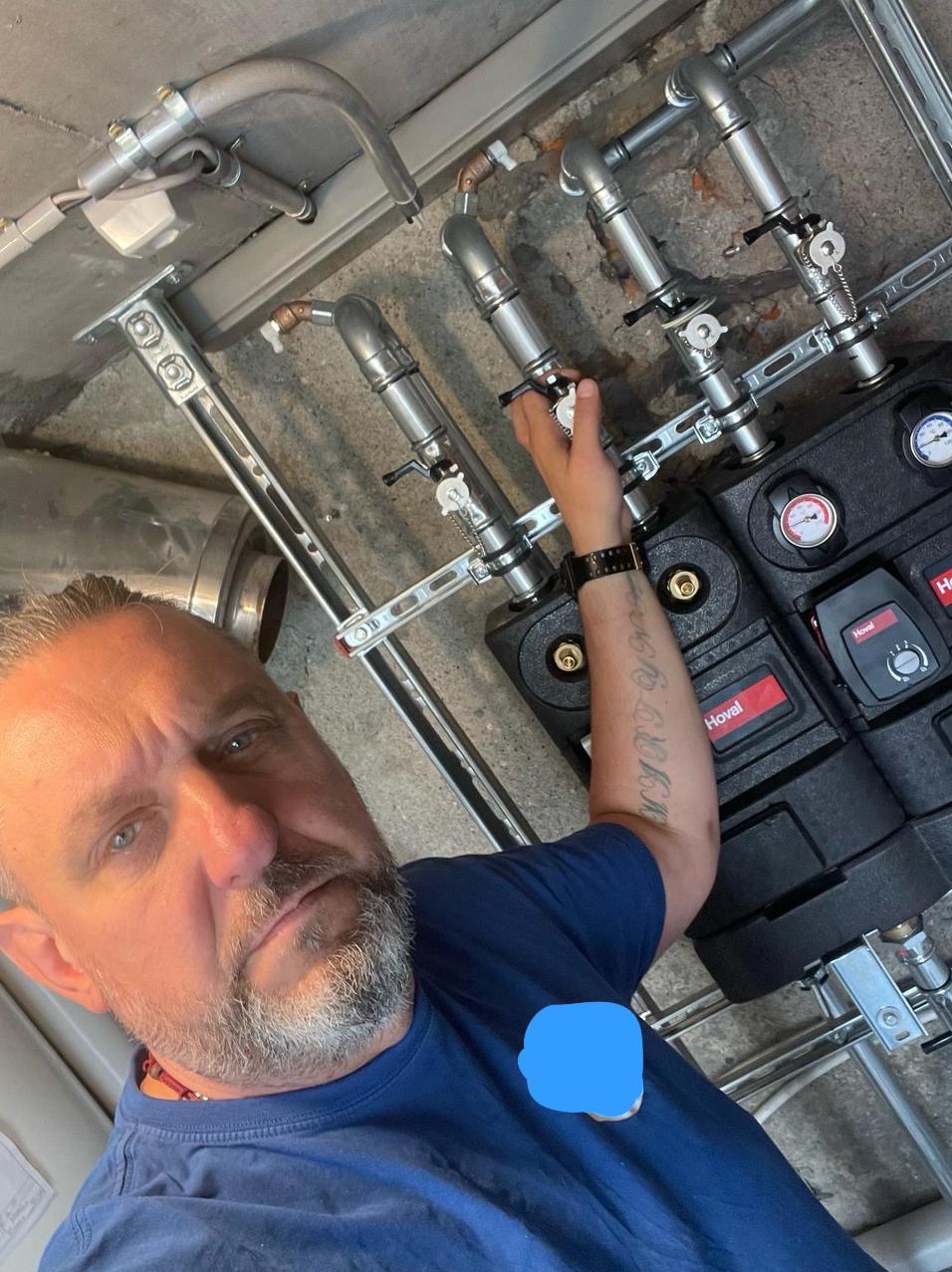 Mężczyzna z tatuażem na ramieniu reguluje zawory przy dwóch czarnych modułach grzewczych Hoval z manometrami, widoczne rury i elementy instalacji grzewczej na ścianie.