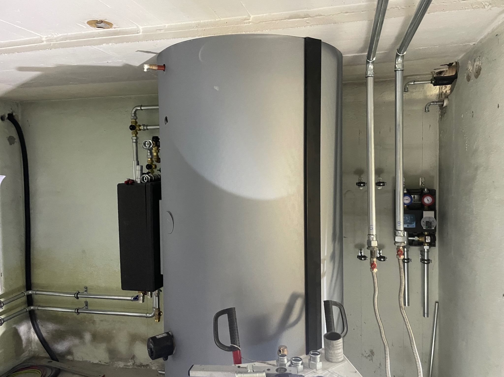 Szary, pionowy zbiornik buforowy ciepła z czarnym panelem sterowania i podłączonymi rurami instalacji grzewczej w surowym pomieszczeniu z betonowymi ścianami i sufitem.