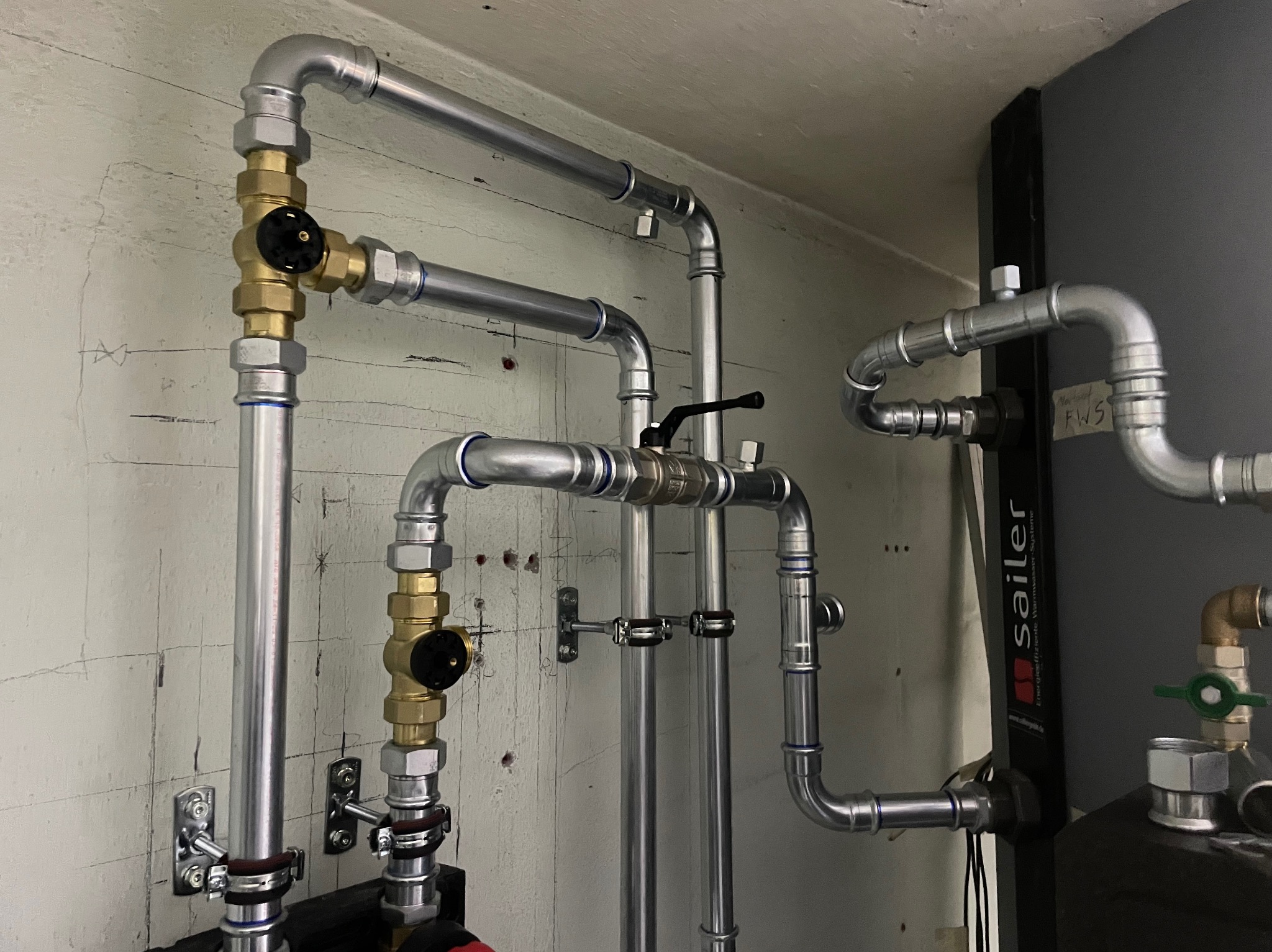 Szczegółowe ujęcie instalacji grzewczej z rurami ze stali nierdzewnej, mosiężnymi zaworami z czarnymi pokrętłami, przymocowanymi do ściany z widocznymi śladami wiercenia, oraz elementami kotła...