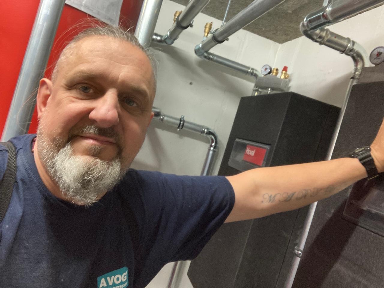 Uśmiechnięty mężczyzna na tle kotłowni z rurami i piecem Hovael. Widoczny fragment instalacji grzewczej i logo firmy na koszulce.