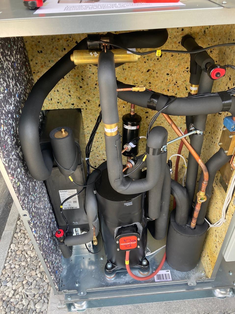 Wnętrze obudowy pompy ciepła z widocznymi elementami instalacji: rurami w izolacji, kompresorem i innymi podzespołami. Widoczne złącza i okablowanie.