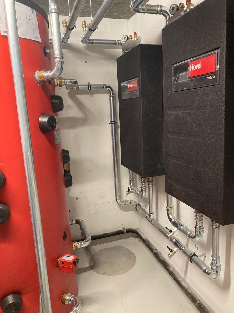 Czerwony zbiornik i dwa czarne piece Hoval z rurami w piwnicy. Instalacja grzewcza z precyzyjnie połączonymi rurami, widoczne zawory i manometry.