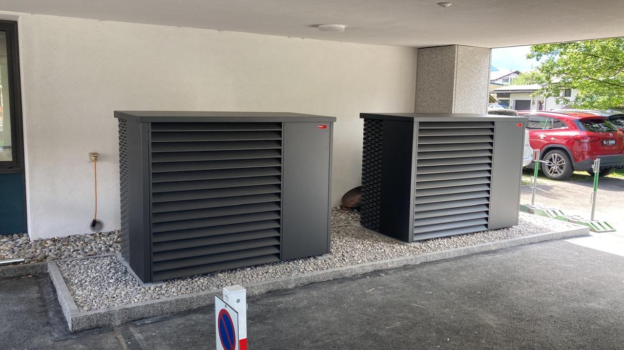 Dwie szare pompy ciepła powietrze-woda, ustawione obok siebie na żwirowym podłożu, pod zadaszeniem. Widoczny fragment parkingu i zaparkowany czerwony samochód.