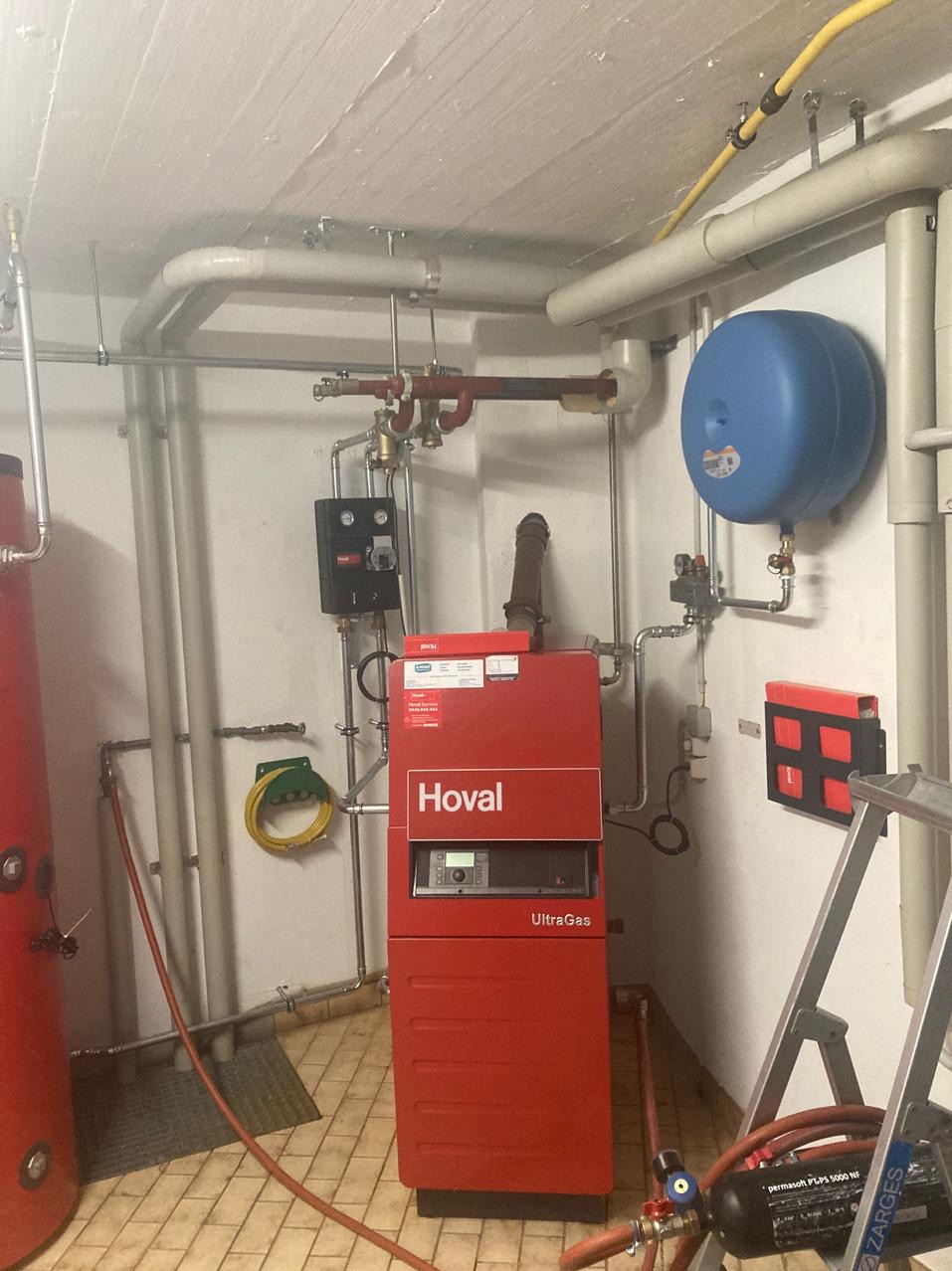 Czerwony piec gazowy Hoval UltraGas w kotłowni, z widocznymi rurami, zbiornikiem wyrównawczym i elementami instalacji. Ujęcie z bliska, ukazujące detale.