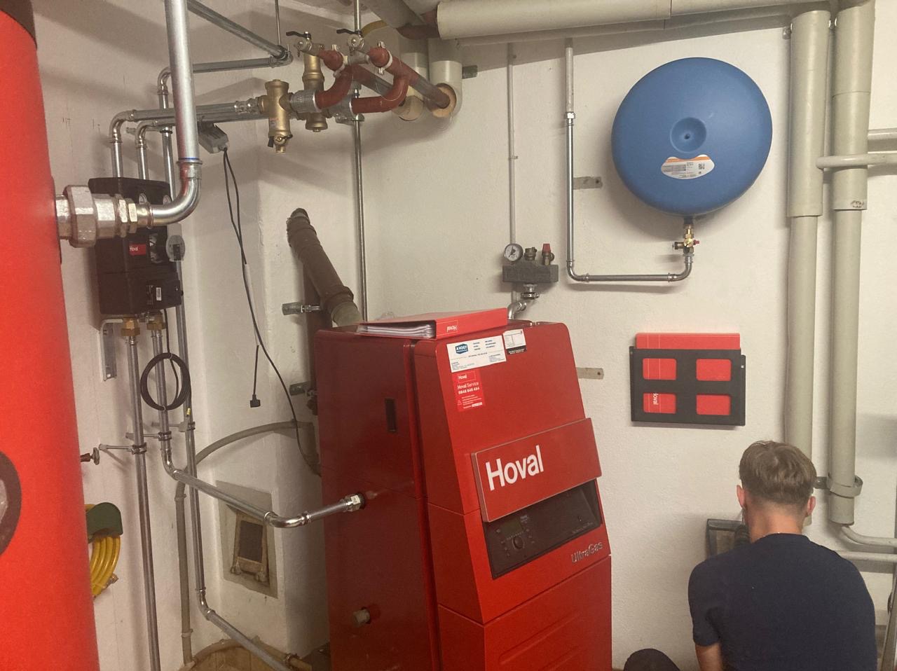 Czerwony piec CO marki Hoval z podłączoną instalacją, widoczny pracownik podczas montażu. Elementy instalacji grzewczej na ścianie i pod sufitem.