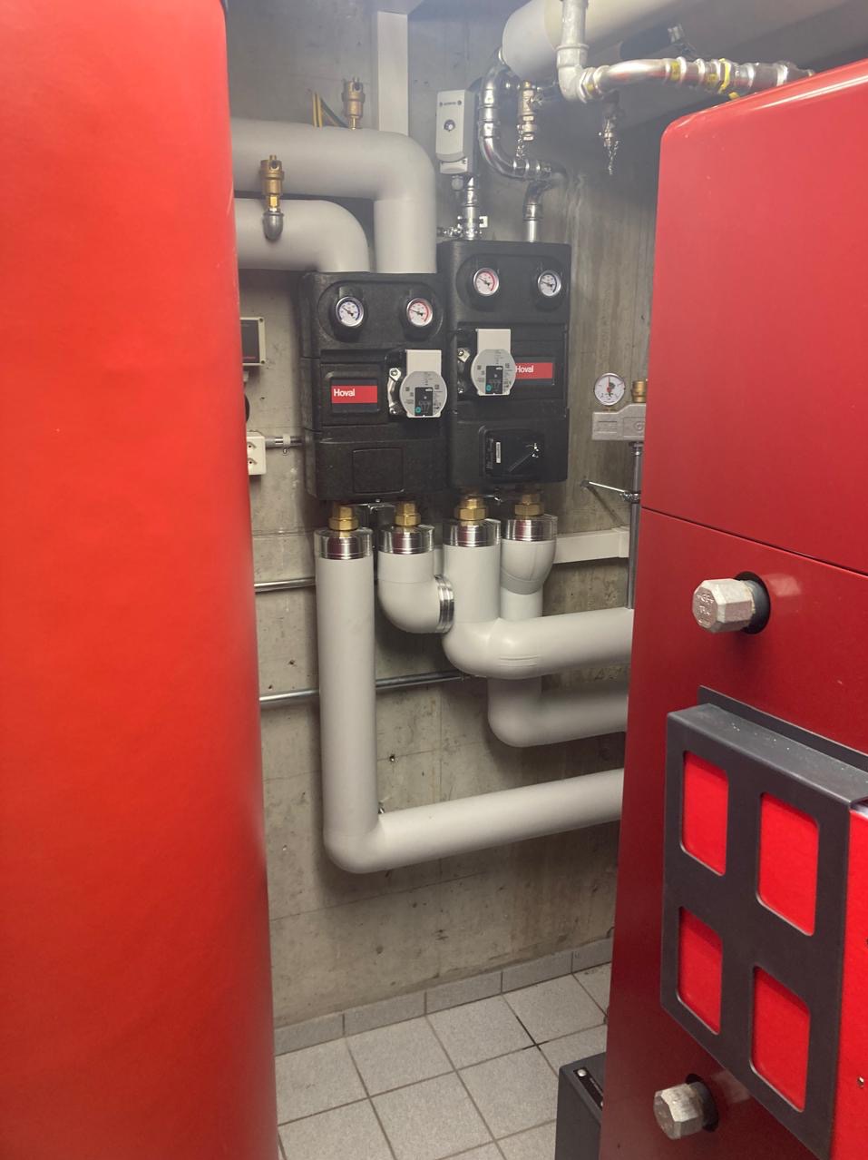 Czerwony piec CO marki Hoval z czarnymi modułami sterującymi i białą instalacją rurową na tle szarej ściany i podłogi z płytek. Widoczne manometry i zawory.