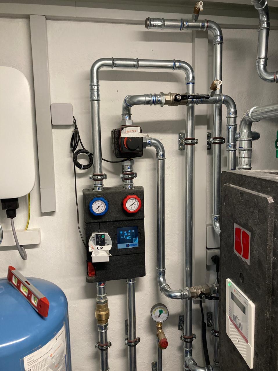 Kompletna instalacja grzewcza z rurami, zaworami, manometrami i sterownikiem, zamontowana na ścianie. Widoczny poziomica na zbiorniku wyrównawczym.