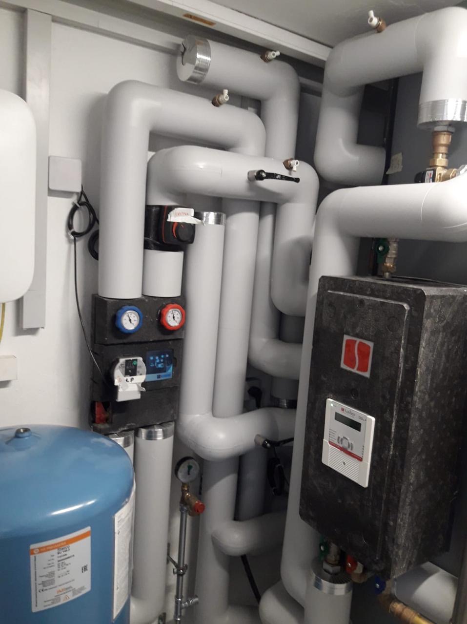 Złożona instalacja grzewcza z rurami w kolorze białym, manometrami i elementami sterującymi. Widoczny zbiornik wyrównawczy w kolorze niebieskim.