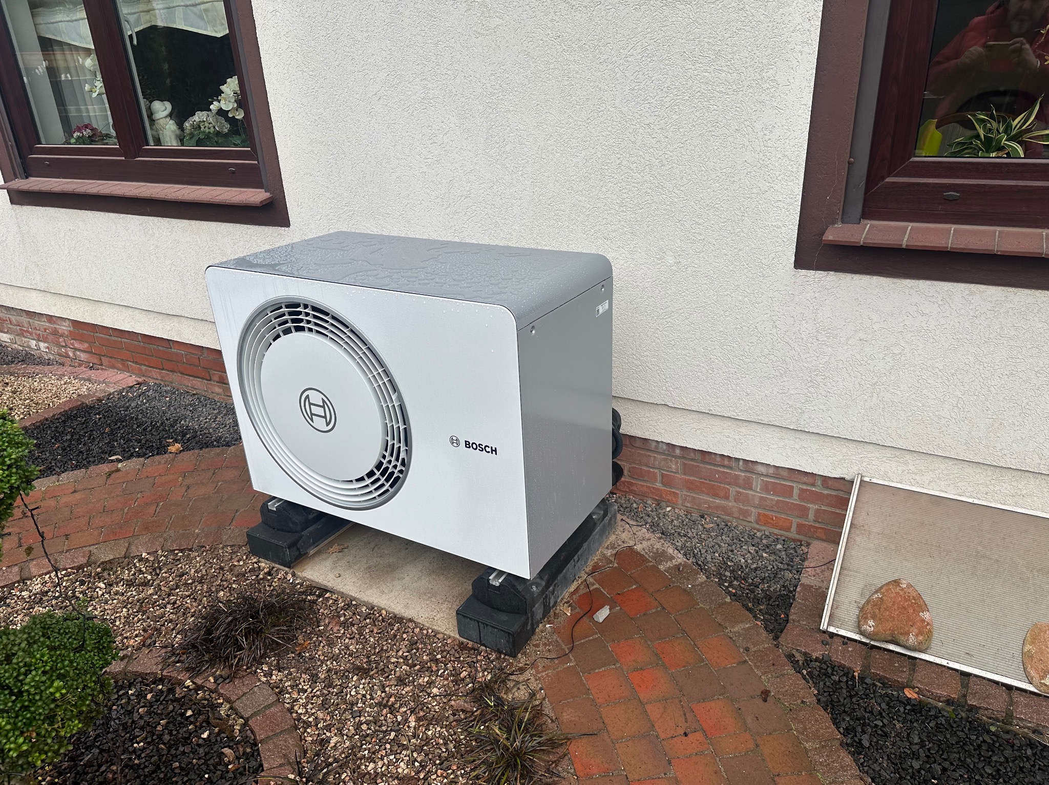 Biała pompa ciepła Bosch na czarnych podkładkach, ustawiona przy ścianie domu z ceglaną podmurówką i dwoma oknami. Drobny deszcz na obudowie. Ogród z kamykami i cegłą.