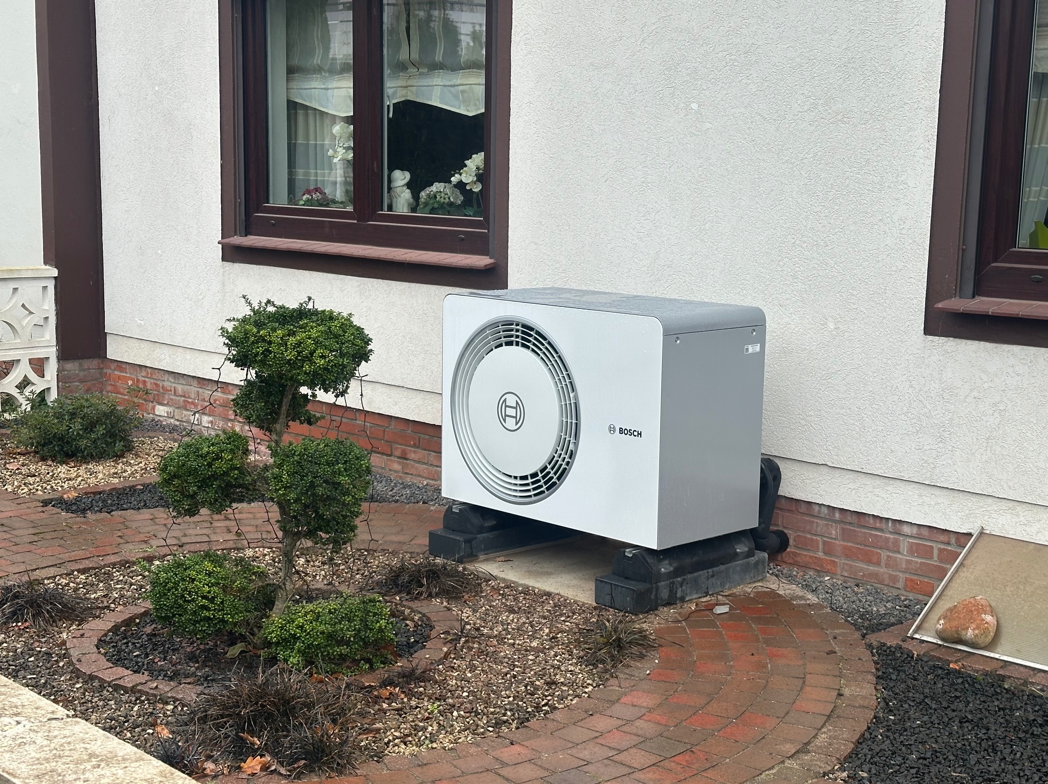 Biała pompa ciepła Bosch na zewnątrz budynku, umieszczona na czarnych podstawach, otoczona ozdobnym ogrodem z krzewami i ścieżką z czerwonej cegły.