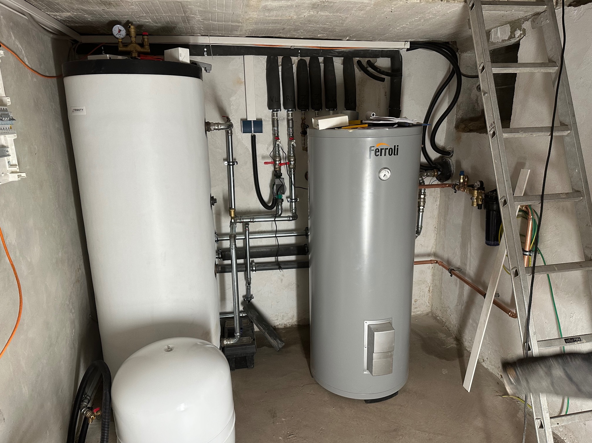 Dwa zbiorniki wody (biały i szary Ferroli) w kotłowni, z widocznymi rurami, zaworami i drabiną. Instalacja w surowym pomieszczeniu z betonową ścianą.