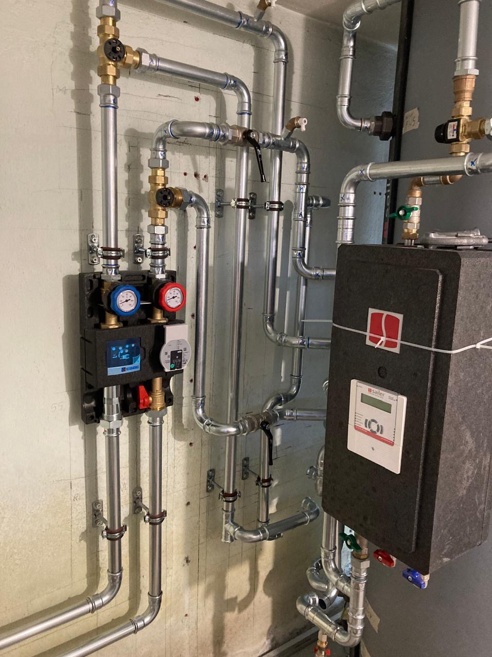 Złożona instalacja grzewcza z rurami, zaworami i czarnym modułem sterującym z wyświetlaczem, zamontowana na surowej ścianie. Widoczne liczne złącza i elementy montażowe.