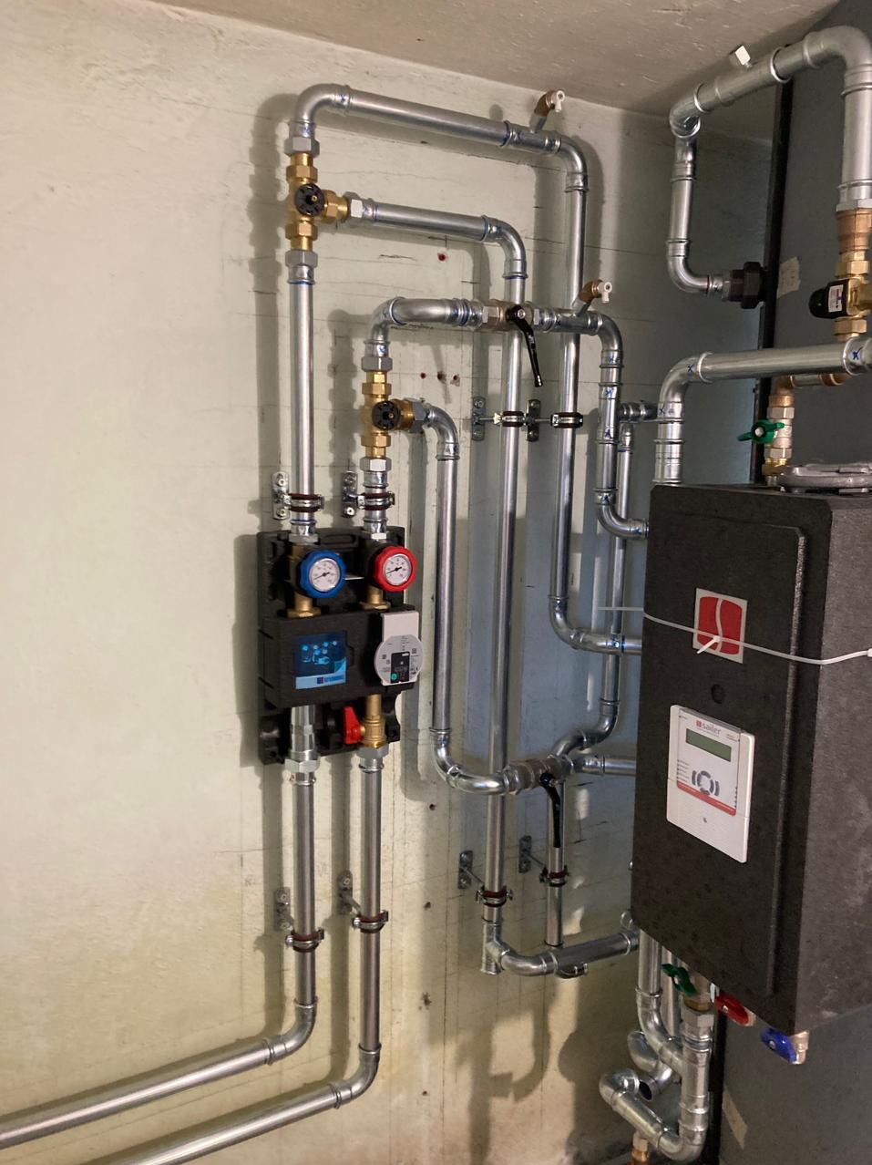 Złożona instalacja grzewcza z rurami, zaworami i czarnym panelem sterującym na tle surowej ściany. Widoczne manometry i liczne złączki.