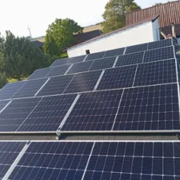 Instalacja paneli fotowoltaicznych na dachu budynku, widok z góry. Na drugim planie budynki gospodarcze i drzewa. Instalator z podniesionymi rękoma.