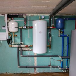 BE-ENERGY - Kompletna instalacja grzewcza z białym zasobnikiem ciepłej wody, niebieskim naczyniem przeponowym i szarymi rurami w piwnicy z surowymi ścianami. Widoczny manometr i zawory.